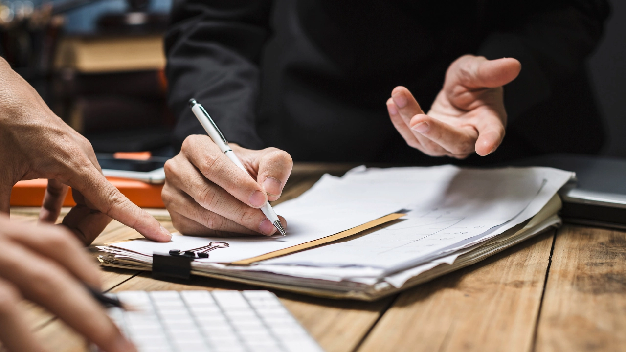 Ein macht Notizen auf Papier auf einem Klemmbrett. Eine andere Person tippt auf einer Tastatur.